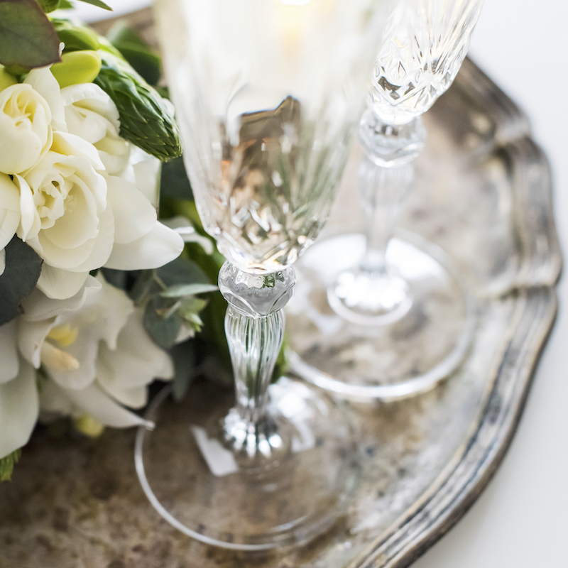 Vintage crystal toasting champagne flutes are staged on an antique silver-plated tray with a bouquet of white flowers.